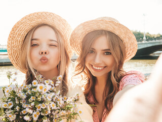 Two young beautiful smiling hipster woman in trendy summer sundress. Sexy carefree women posing on the street background in hats. Positive models at embankment at sunset. Taking selfie.Holding flowers