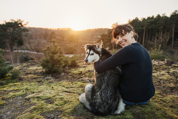 Person and dog are having fun and looking at the camera. The woman and the husky enjoy the view in a beautiful landscape. Hugging at sunset.