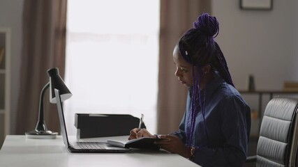 adult black female student is learning online, viewing lecture on laptop screen and making notes in pad, sitting at table in room of apartment