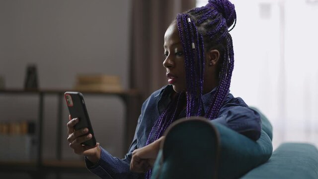 black woman is sitting in living room of her apartment and listening to speaker of working video chat in smartphone, looking at screen
