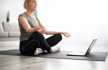 Balanced mature woman meditating with closed eyes in front of laptop at home, selective focus