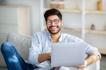 Freelance career. Happy young arab man sitting on sofa and working on laptop, smiling to camera