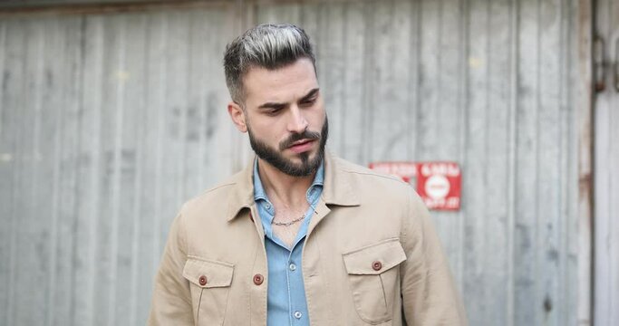 cool bearded man in denim shirt adjusting beige jacket, looking both sides, touching beard and rubbing palms, looking over shoulder and walking in front of metal garage outside