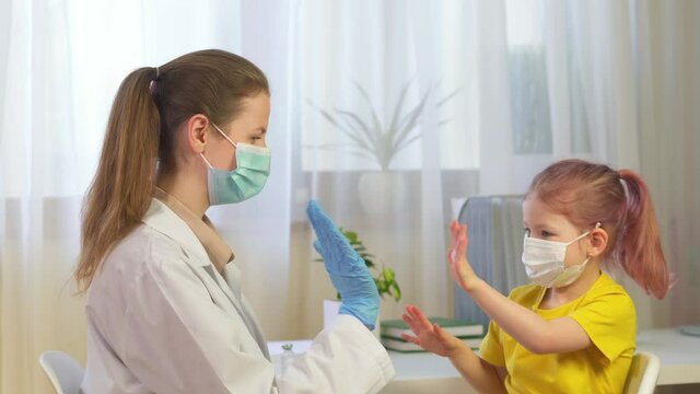 doctor in mask dance and have fun with preschool patient in clinic. High five child and a doctor. The girl gives beats off the woman's class to pediatrician. Quarantine covid in masks and gloves.