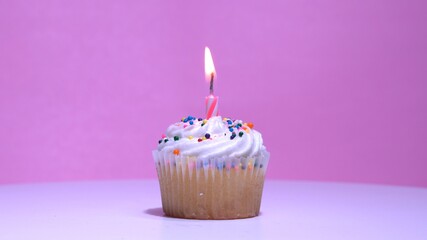 Cupcake with burning festive candle at white cream and sugar icing on pink background, macro shallow depth of field view. Homemade vanilla cup cake with buttercream frosting close up.