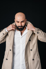 A young bearded man stands and adjusts his raincoat against a black background.