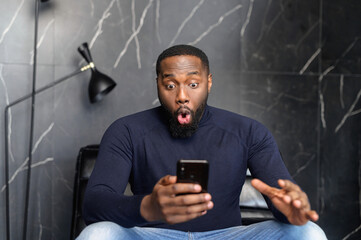 Astonished happy biracial man holding phone, looking at cellphone, read good news in sms, sit on...