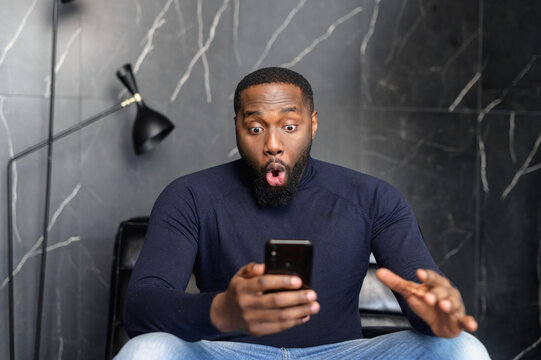 Astonished happy biracial man holding phone, looking at cellphone, read good news in sms, sit on armchair, amazed black guy winner excited by mobile app, win scream with joy, celebrate his victory