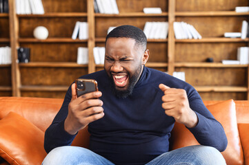 Excited black multiracial man sitting on the brown sofa looking at the phone and screaming, surprised emotional lucky man with wide opened mouth, rejoicing at good message, news, won the lottery