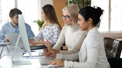 Focused multiracial businesspeople sit at desk in office look at PC screen brainstorm discussing company financial ideas or project. Serious diverse female employees cooperate using computer together.