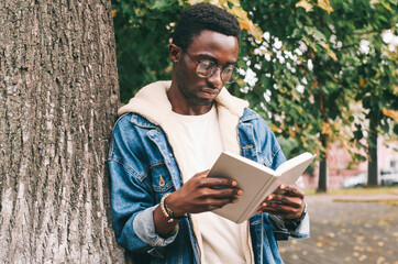 Fototapeta premium Portrait of young african man student reading a book wearing an eyeglasses in autumn city park