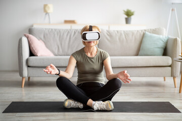 Full length of senior lady in VR glasses practicing yoga or meditating in lotus pose at living room