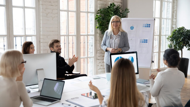 Diverse businesspeople applaud clap hands thanking middle-aged successful female coach or trainer for presentation. Happy excited employees show acknowledgement for businesswoman at briefing.