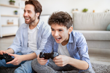 Quarantine entertainment. Overjoyed dad and son competing for win on gamepad at home, spending...