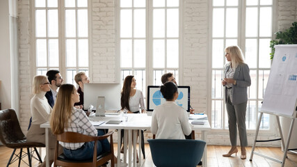 Middle-aged Caucasian businesswoman hold lead meeting present business project on whiteboard for colleagues in office. Successful female coach make flip chart presentation for employee at briefing.