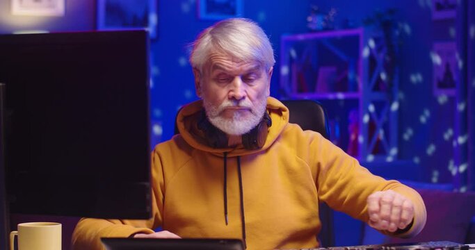 Close up portrait of handsome joyful senior man DJ playing on equipment at home in colorful neon lights recording music working and typing on computer. Old male sound beat maker writing creative music
