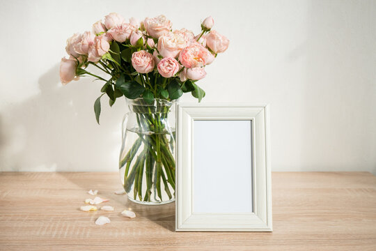 Portrait white picture frame mockup on wooden table. Modern glass vase with roses. White wall background. Scandinavian interior. 