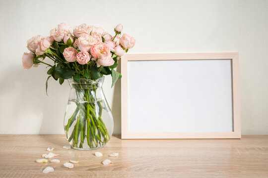 Portrait white picture frame mockup on wooden table. Modern glass vase with roses. White wall background. Scandinavian interior. 
