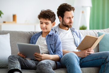 No communication in family. Father reading book while his son using digital tablet, sitting together on sofa