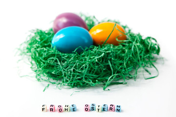 Colorful easter eggs in a nest of green grass on white background and cubic letters saying Happy Easter in German.