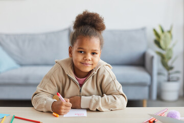 Front view portrait of cute African-American girl drawing pictures and smiling at camera while sitting at desk in cozy home interior, copy space