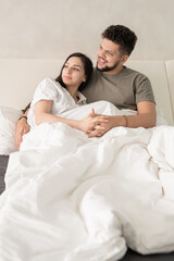 Young affectionate restful couple relaxing on comfortable bed in the morning and looking aside while discussing something pleasant