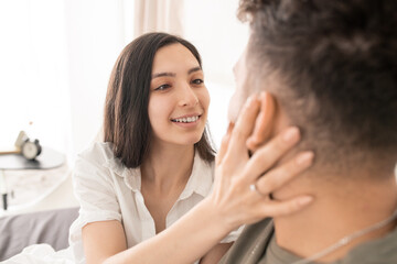 Happy young brunette woman looking at her husband eyes with kind smile while touching his face and wishing him good morning after sleep