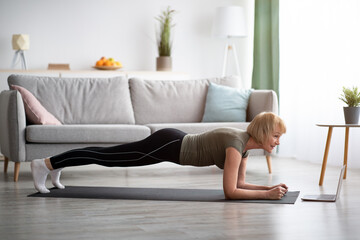 Side view of mature woman standing in elbow plank,doing yoga asana, training to online sports video at home, copy space