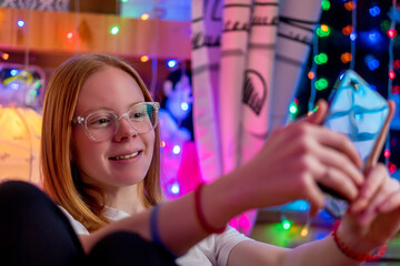 cute teenage girl wearing glasses makes selfie on smartphone on blurry garland background