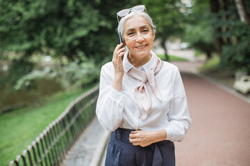 Smiling senior woman talking on mobile while relaxing at green park. Happy grey-haired lady enjoying conversation while walking on fresh air.