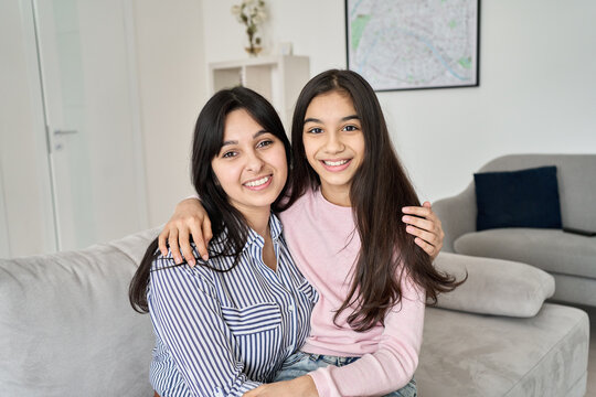 Happy teen child daughter hugging young indian mother at home, portrait. Smiling ethnic family mom with teenage kid girl embracing, bonding together looking at camera sitting on couch.