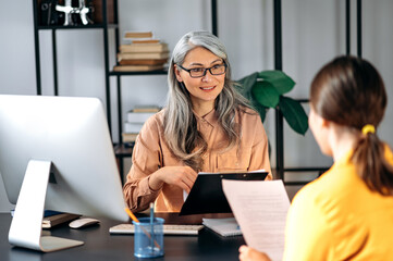 Intelligent confident influential gray-haired female asian wearing glasses, business lady, HR manager, talking to job candidate in modern office, holds and studies resume, smiling friendly - obrazy, fototapety, plakaty