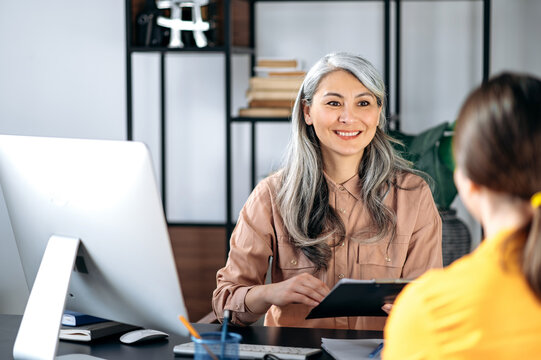 Intelligent Confident Influential Gray-haired Female Asian Wearing Glasses, Business Lady, HR Manager, Talking To Job Candidate In Modern Office, Holds And Studies Resume, Smiling Friendly