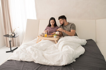 Obraz na płótnie Canvas Young affectionate couple in t-shirts sitting in bed and having tasty breakfast while female spreading jam on slice of wheat bread