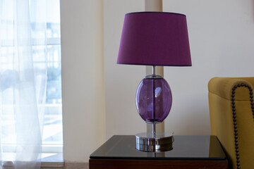Purple lamp with a shade stands on table near a window. Stylish table lamp