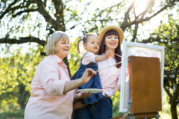 Fototapeta premium Easel, canvas, brushes and paints. Family leisure. Close up bottom view of three generation family, grandmother, mother and little girl artist, painting a picture on canvas outdoors together.