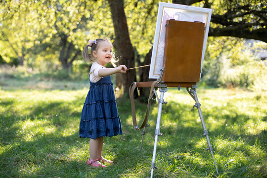 Creative hobby and leisure activity. Little talented artist preschool girl, having fun while painting a picture on easel outdoors at nature in sunny garden at bright summer day.