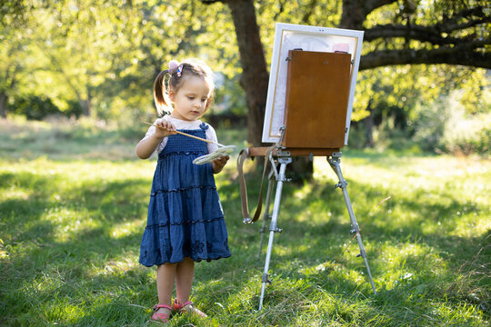Painting on easel outdoors in the nature. Little cute talented excited kid girl artist, painting a picture on canvas, holding palette and brush and choosing a paint color.