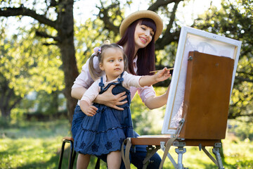 Close up of young charming mother with her little daughter, wearing jeans clothes, painting together on easel outdoors in bright summer garden. Mother's Day Concept.