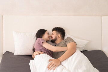 Happy young affectionate couple in t-shirts lying in embrace under white blanket on double bed and touching by noses and keeping eyes closed