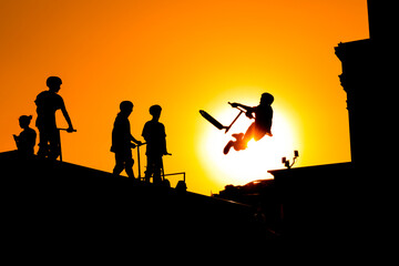 Unrecognizable teenage boy silhouette showing high jump tricks on scooter against orange sunsetwarm...