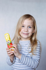 Blonde girl in striped jacket on a light background holds a yellow hare in front of her face smiling