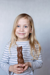 Blonde girl in striped jacket on a light background bites chocolate hare in the face smiles look to the side