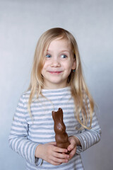 Blonde girl in striped jacket on a light background bites chocolate hare in front of her face smiles