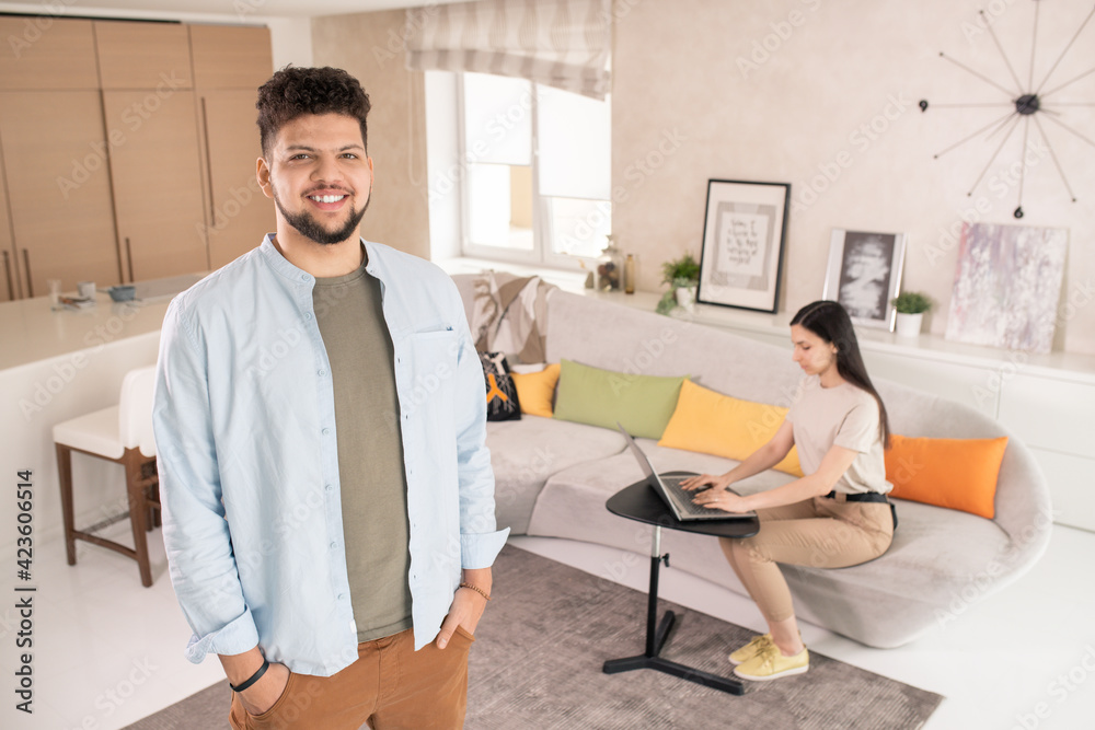 Poster young cheerful man in casualwear looking at you while standing against his wife sitting on couch and