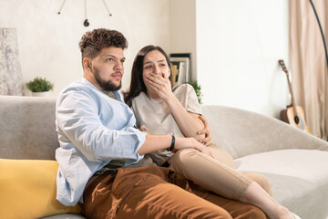 Young scared brunette woman covering mouth by hand while sitting on couch close to her husband and both watching horror movie