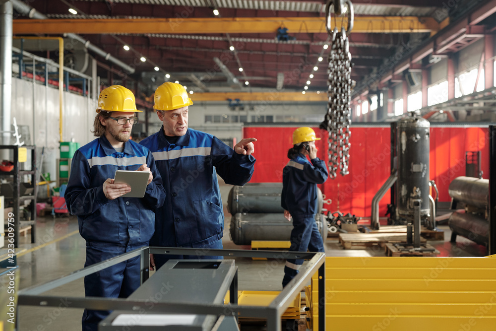 Poster two engineers in workwear standing in large workshop or plant and discussing new industrial equipmen