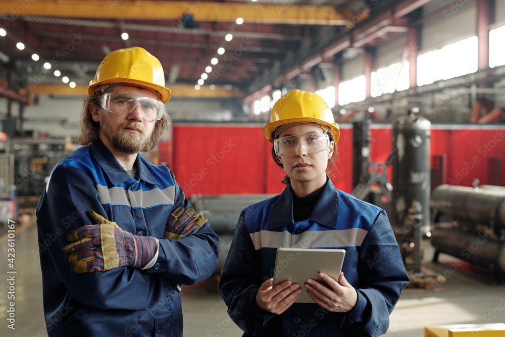 Poster two young serious engineers in protective workwear looking at you while standing in front of camera