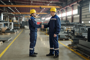 Side view of two male engineers in protective hardhats and workwear greeting one another by handshake in the morning in workshop