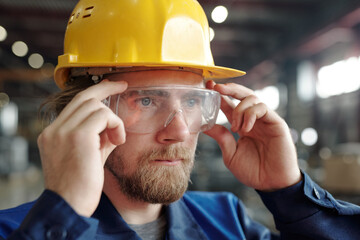 Young serious male engineer or factory worker in protective helmet and eyeglasses standing in front...